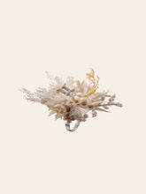 The Gemini Flower Ring by CAMELLIA is handcrafted in New York, featuring a mix of beige and brown dried flowers on an adjustable circular band. This floral ring captures the timeless elegance of nature set against a plain background.